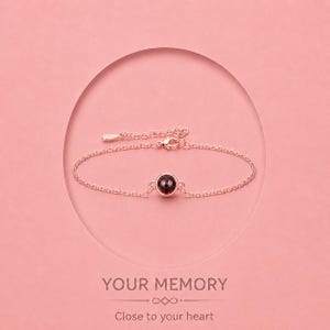 Peut inclure: Bracelet en or rose avec une pierre précieuse ronde et rouge foncé. Le bracelet est présenté sur un fond rose avec le texte "YOUR MEMORY" et "Close to your heart". Le bracelet a une chaîne réglable.