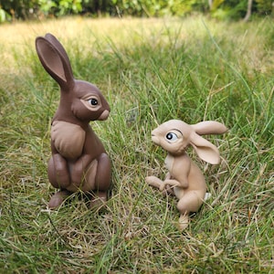 Könnte beinhalten: Zwei Kaninchenfiguren auf einer Grasfläche. Das größere Kaninchen ist dunkelbraun mit einer helleren beigen Brust und langen Ohren. Das kleinere Kaninchen ist hellbeige mit großen Augen und langen Ohren. Beide Figuren haben glatte Oberflächen und detaillierte Gesichtszüge.