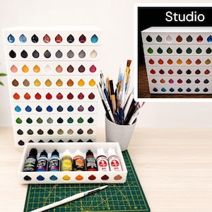 May include: A white modular paint storage rack with multiple horizontal tiers featuring teardrop-shaped cutouts for paint bottles. The system includes a separate tray holding several small plastic paint bottles and a cup filled with various paintbrushes.