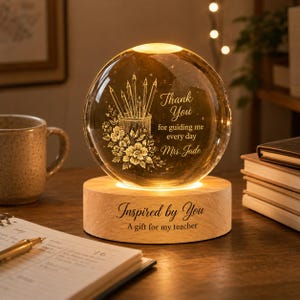 Può includere: Una sfera di cristallo illuminata su una base di legno. La sfera presenta un disegno inciso di matite, fiori e il testo "Thank You for guiding me every day Mrs. Jade." La base recita "Inspired by You, A gift for my teacher."