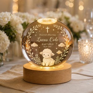 May include: A personalized glass sphere with a wooden base, illuminated from within. The sphere features the text "Heaven's Blessing" and "Lucas Cole BAPTIZED 08.03.2026", along with a lamb illustration and decorative elements. The sphere is lit with a warm glow.