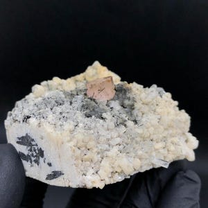 May include: A mineral specimen featuring a single, translucent, pale pink cubic crystal centered atop a cluster of small, off-white, granular crystals. The base includes patches of dark gray, metallic-looking mineral inclusions embedded within.