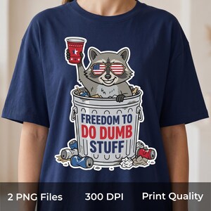 May include: A navy blue t-shirt featuring a graphic of a raccoon wearing American flag sunglasses, sitting in a trash can, holding a red cup, and the text FREEDOM TO DO DUMB STUFF. The design includes scattered trash items like cans and paper.