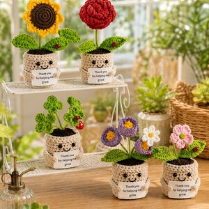 Op de afbeelding: Gehaakte bloempotten met lachende gezichten, elk met een bordje met de tekst "Thank you for helping me grow". De bloemen zijn in verschillende kleuren, waaronder geel, rood en paars. De potten zijn beige en staan op een wit rek.