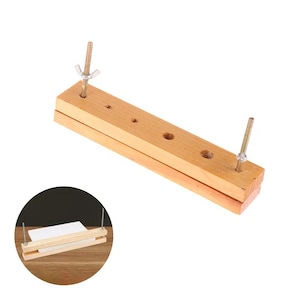 May include: A light-coloured wooden bookbinding press consisting of two rectangular blocks held together by two threaded metal rods with wing nuts. The top block features several circular holes, and the device is shown clamping a white paper stack.