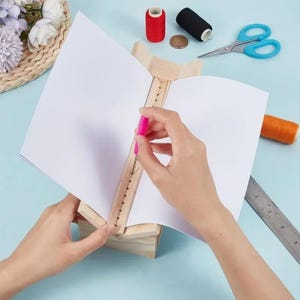 Puede incluir: Una cuna de encuadernación de madera con una ranura de alineación central que sostiene una pila de papel blanco. Un punzón rosa está colocado en la ranura, rodeado de suministros de costura que incluyen carretes de hilo rojo y negro, tijeras y una regla.