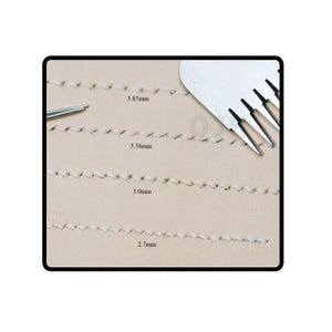 May include: A light beige leather surface featuring four parallel rows of white stitching. Each row is labeled with its specific spacing measurement: 3.85mm, 3.38mm, 3.0mm, and 2.7mm. A silver metal stitching chisel tool is positioned at the top right.