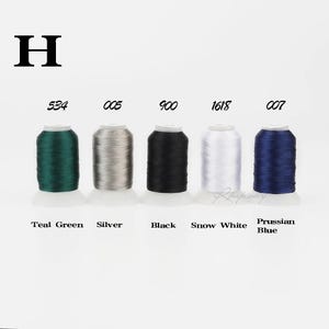 Puede incluir: Cinco carretes de hilo de bordar etiquetados con la letra H, en colores 534 Teal Green, 005 Silver, 900 Black, 1618 Snow White y 007 Prussian Blue. Cada carrete es cilíndrico con una base blanca y muestra claramente su código y nombre.