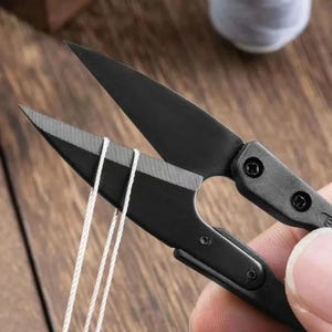 Puede incluir: Unas tijeras cortahilos de metal negro mate con hojas afiladas y puntiagudas. Las tijeras se muestran en posición abierta, con tres hebras de hilo blanco fino colocadas entre las hojas, y cuentan con tornillos visibles en el mango.