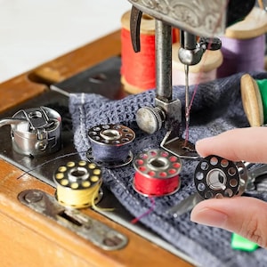 May include: A vintage sewing machine with a metal presser foot and needle, featuring several metal bobbins wound with yellow, red, and blue thread. A dark blue textured fabric sits under the needle, with additional spools of thread visible.