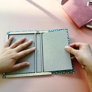 May include: A light-colored wooden bookbinding jig with the text 4x6 printed on the top edge. The jig is positioned over a piece of blue paper with white polka dots and gray cardboard panels, used for assembling a 4x6 inch book cover.
