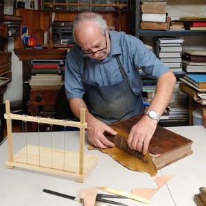 May include: A wooden bookbinding sewing frame with five vertical metal wires and adjustable wing nuts, positioned on a white surface next to a large leather-bound book, various leather scraps, and metal bookbinding tools.