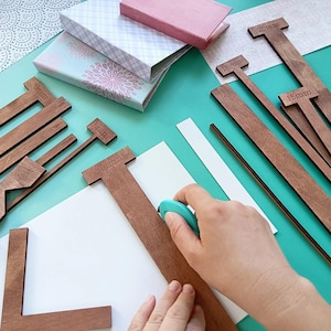 May include: A collection of brown wooden bookbinding guides and rulers with engraved measurements including 5mm, 10mm, 15mm, 20mm, 25mm, and 30mm. A turquoise handheld craft tool is positioned against a 30mm guide on a white sheet of paper.