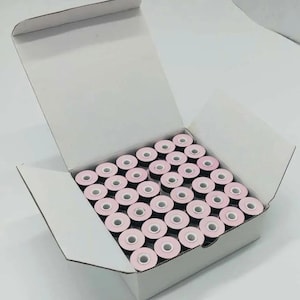 Puede incluir: Una caja de cartón blanca llena con una cuadrícula de bobinas para máquina de coser. Cada bobina presenta un hilo oscuro enrollado alrededor de un núcleo central con un centro circular de color rosa claro. La caja está abierta y organizada.