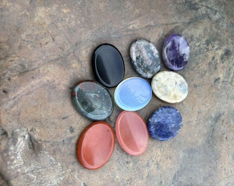 May include: A collection of eight smooth, oval-shaped polished stones in various colours including solid black, translucent light blue, deep red, mottled grey, purple, cream, and dark blue with white veining, arranged in a cluster.