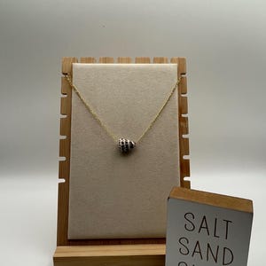 Puede incluir: Un collar de cadena en tono dorado con un colgante de concha en forma de cono con un patrón de manchas blancas y marrones. El collar se exhibe en un soporte de joyería beige claro junto a un pequeño letrero de madera con el texto SALT SAND SURF.