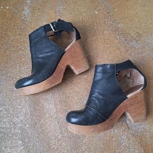 May include: A pair of black leather ankle boots with wooden heels and a cut-out design. The boots have a buckle closure and a pointed toe.