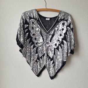 Può includere: Un top nero e argento con paillettes e un design a farfalla. Il top ha una scollatura a V e maniche corte.