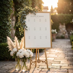 May include: A gold framed seating chart with the names "Olivia & Henry" at the top, displayed on a gold easel. The chart is decorated with greenery. Two vases of white flowers and pampas grass are in front of the easel.
