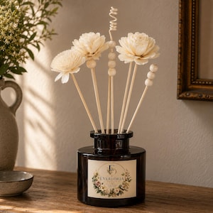 Op de afbeelding: Een donkerbruine glazen diffuserfles met een label met de tekst "EVERLORIA DECOR". De fles bevat lichtgekleurde bloemvormige rietjes en houten kralen. De opstelling staat op een houten oppervlak.