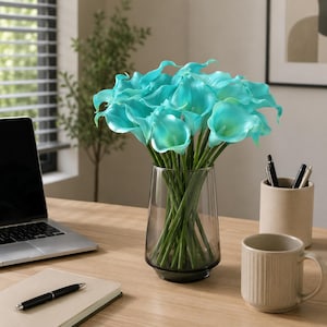 Puede incluir: Un ramo de lirios cala de color turquesa vibrante con pétalos curvos y lisos y tallos verdes largos, dispuesto en un jarrón de vidrio transparente y cónico. Las flores presentan un tono azul verdoso brillante y saturado.