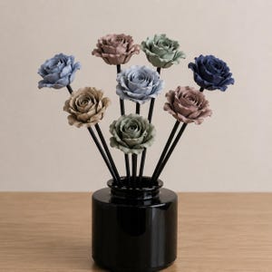 Puede incluir: Un jarrón negro cilíndrico contiene una colección de rosas artificiales en varios colores apagados, incluyendo azul, rosa y verde. Las rosas están montadas sobre tallos negros, creando un arreglo floral decorativo. El jarrón está sobre una superficie de madera.