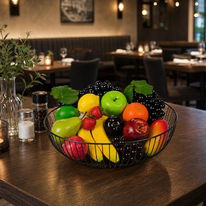 Peut inclure: Une corbeille à fruits en fil métallique noir remplie de divers fruits artificiels, notamment une pomme verte, un citron jaune, une pomme rouge, une orange, une mangue, une poire, un citron vert, des fraises et des grappes de raisins violets.