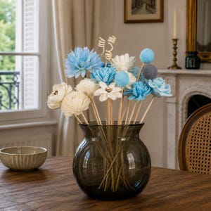 Puede incluir: Un arreglo decorativo de flores artificiales en un jarrón de cristal oscuro. Las flores son en tonos azules, blancos y crema, algunas parecen ser de madera. El jarrón está sobre una mesa de madera, con un cuenco pequeño en primer plano.
