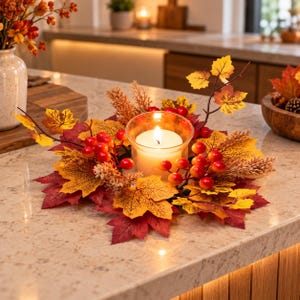 Peut inclure: Un anneau de bougie composé de feuillage d'automne artificiel, incluant des feuilles d'érable rouges et jaunes, des grappes de petites baies rouges et des brins beiges texturés. Un photophore en verre clair contient une bougie blanche allumée.