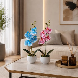 May include: Two artificial orchid plants in white ceramic pots. One plant features vibrant blue petals with white speckles, while the other displays pink petals with white speckles. Both orchids have green stems, dark green leaves, and small buds.