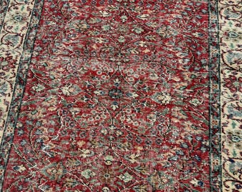 May include: A rectangular area rug featuring a deep red central field with an intricate floral and vine pattern in shades of blue, cream, and green. The rug is framed by a wide, ornate border with repeating botanical motifs on a light background.
