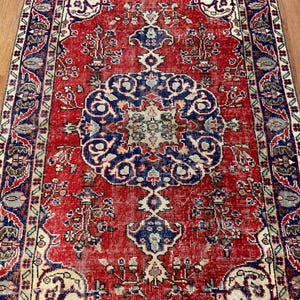Pu&ograve; includere: Un tappeto rettangolare caratterizzato da un campo rosso vivace con un grande medaglione centrale blu scuro. Il design presenta intricati motivi floreali, schemi simmetrici e un bordo decorativo nei toni del blu, crema e verde spento.