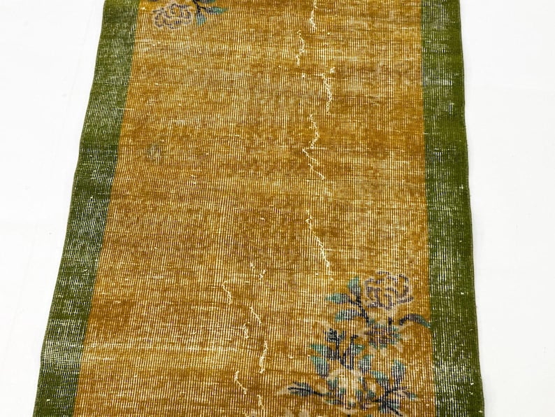 May include: A rectangular runner rug featuring a central mustard yellow field with a distressed texture and a faded floral motif in blue and white. The rug is framed by solid green borders on the left and right sides, showcasing a woven construction.
