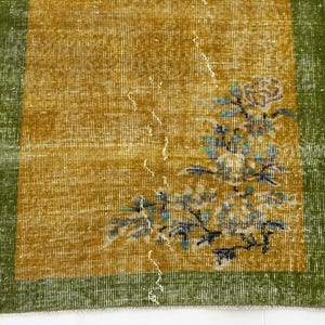 May include: A rectangular rug featuring a mustard yellow centre field with a distressed texture and a floral motif in shades of blue, white, and green. The rug is framed by a solid dark green border on all sides, showcasing a woven textile construction.