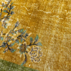 May include: A woven textile rug featuring a mustard yellow field with a distressed, faded appearance. A floral pattern in shades of teal, white, and dark grey branches across the surface. A solid green border runs along the bottom edge.