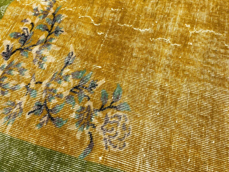 May include: A woven textile rug featuring a mustard yellow field with a distressed, faded appearance. A floral pattern in shades of teal, white, and dark grey branches across the surface. A solid green border runs along the bottom edge.