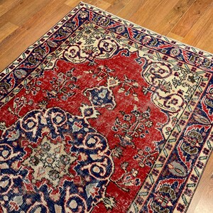 Pu&ograve; includere: Un tappeto rettangolare caratterizzato da un vivace campo centrale rosso con un grande e intricato medaglione blu e crema. Il bordo presenta un motivo ripetitivo di disegni floreali blu, rossi e crema, creando un design simmetrico.