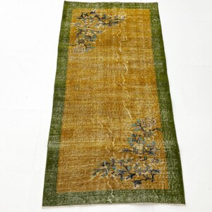 May include: A rectangular runner rug featuring a mustard yellow centre field with a distressed texture. The rug is framed by a wide, dark green border and decorated with floral motifs in shades of blue, white, and brown at the corners.