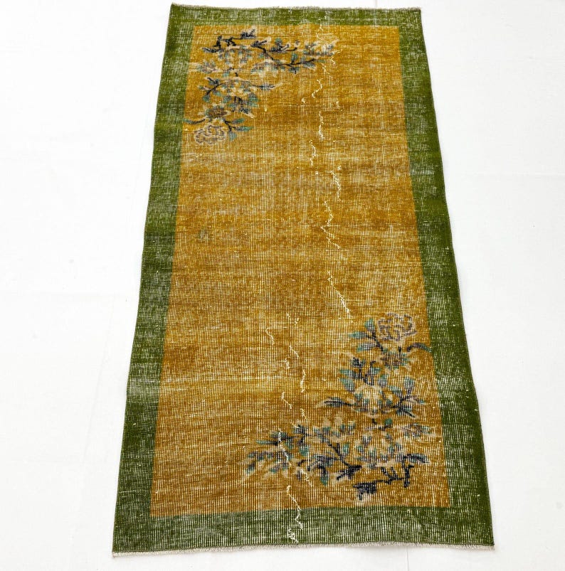 May include: A rectangular runner rug featuring a mustard yellow centre field with a distressed texture. The rug is framed by a wide, dark green border and decorated with floral motifs in shades of blue, white, and brown at the corners.