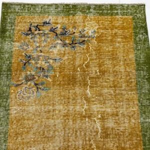 May include: A rectangular area rug featuring a mustard yellow centre field and a wide olive green border. The rug displays a floral branch motif in the upper left corner with blue, black and white details, showing a distressed, worn texture.