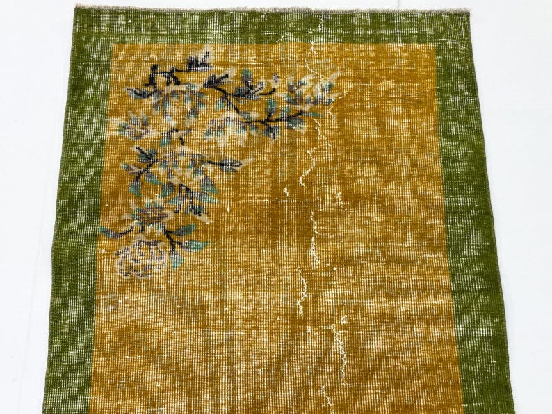 May include: A rectangular area rug featuring a mustard yellow centre field and a wide olive green border. The rug displays a floral branch motif in the upper left corner with blue, black and white details, showing a distressed, worn texture.