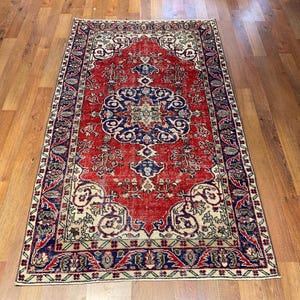 Pu&ograve; includere: Un tappeto rettangolare caratterizzato da un campo centrale rosso vivace con un grande e intricato medaglione blu e crema. Il tappeto &egrave; incorniciato da un ampio bordo blu scuro decorato con motivi floreali e di viti ripetuti nei toni del rosso, crema e verde.