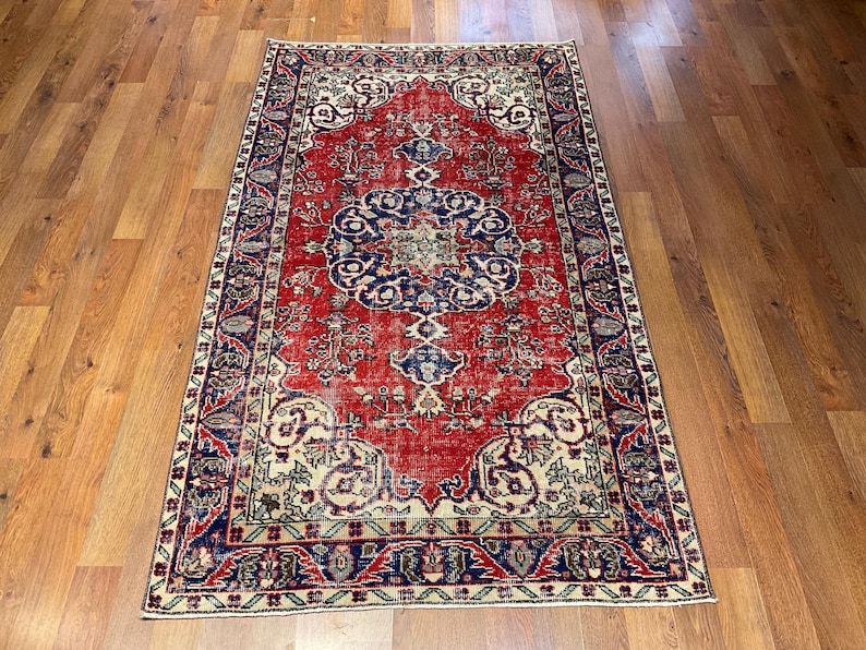 Pu&ograve; includere: Un tappeto rettangolare caratterizzato da un campo centrale rosso vivace con un grande e intricato medaglione blu e crema. Il tappeto &egrave; incorniciato da un ampio bordo blu scuro decorato con motivi floreali e di viti ripetuti nei toni del rosso, crema e verde.