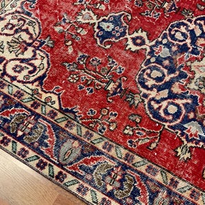 Pu&ograve; includere: Un tappeto rettangolare con uno sfondo rosso vivace e intricati motivi floreali e geometrici in blu profondo, crema e verde chiaro. Il design include un motivo a medaglione centrale e un bordo dettagliato con forme ripetitive.