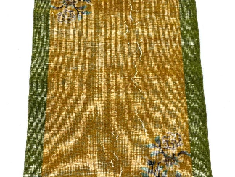 May include: A rectangular runner rug featuring a central mustard yellow field with a distressed texture, flanked by solid green borders on the left and right sides. The rug displays floral motifs in blue and white at the top and bottom corners.