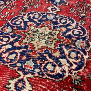 Pu&ograve; includere: Un tappeto rettangolare con uno sfondo rosso vivace e un grande medaglione centrale circolare in blu profondo, crema e azzurro. Il design presenta intricati motivi floreali e geometrici su tutto il campo e lungo i bordi.