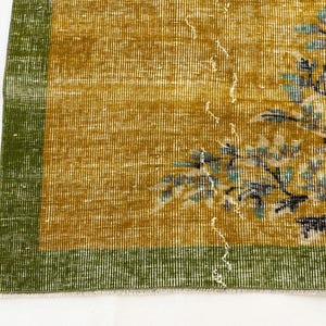May include: A rectangular rug featuring a mustard yellow field with a green border. The rug displays a floral pattern in shades of light blue, white, and dark brown. The weave shows a distressed texture with visible white thread details throughout.