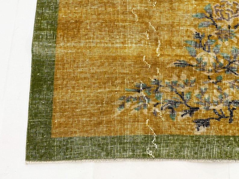May include: A rectangular rug featuring a mustard yellow field with a green border. The rug displays a floral pattern in shades of light blue, white, and dark brown. The weave shows a distressed texture with visible white thread details throughout.
