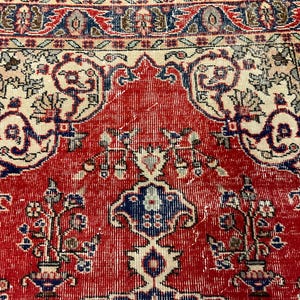 Pu&ograve; includere: Un tappeto tessuto caratterizzato da un campo centrale rosso con un motivo floreale simmetrico in blu, crema e rosso scuro. Il design include vasi stilizzati, viticci e un bordo decorativo con motivi geometrici nei toni del blu e del crema.