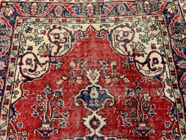 Pu&ograve; includere: Un tappeto tessuto caratterizzato da un campo centrale rosso con un motivo floreale simmetrico in blu, crema e rosso scuro. Il design include vasi stilizzati, viticci e un bordo decorativo con motivi geometrici nei toni del blu e del crema.